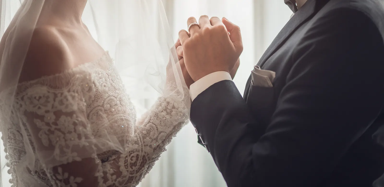A bride and groom holding hands, her lace sleeve against his dark suit